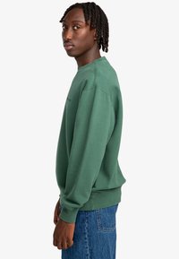 Grüner Baumwollpullover mit Rundhalsausschnitt und gerippten Bündchen, in einem lässigen Schnitt. Kombiniert mit blauen Denim-Jeans.