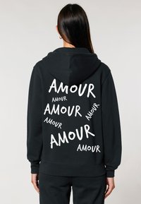 Čierna mikina s kapucňou, ktorá na zadnej strane nesie biele písmo s nápisom "AMOUR" v rôznych veľkostiach. Mäkká tkanina, voľný strih a kapucňa so šnúrkou.