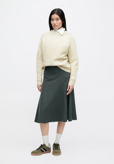 Μπεζ μάλλινο πουλόβερ με χαλαρή εφαρμογή, φορεμένο πάνω από μια σκούρα πράσινη φούστα. Το μοντέλο φοράει πράσινα αθλητικά παπούτσια με μαύρες ρίγες και λευκές κάλτσες.