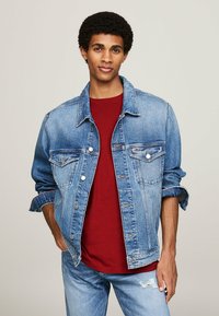 Chaqueta de mezclilla azul clara con corte relajado, dos bolsillos en el pecho, herrajes plateados y mangas enrolladas, llevada sobre una camiseta roja.