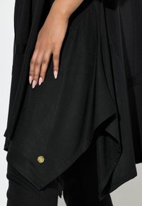 Käsi, jolla on pitkät, vaaleanpunaiset kynnet, lepää kerrostetun mustan epäsymmetrisen kankaan päällä lähellä helmaa, jossa on pieni pyöreä kultainen merkki.