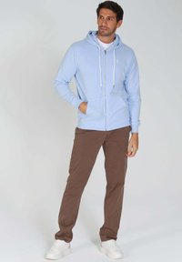 Haut de survêtement zippé bleu clair avec cordons, doté d'une poche frontale, associé à un pantalon marron et des baskets blanches. Texture lisse, coupe décontractée.