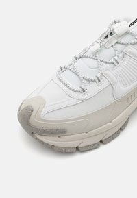 Vit sneaker med texturerade paneler, med elastiska snören, en vadderad tunga och en tjock, prickig sula för stabilitet och komfort.