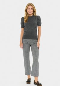 Femeie stând în picioare, zâmbind, îmbrăcată cu un top gri cu mânecă scurtă, pantaloni gri cu model și pantofi din piele maro pe un fundal alb simplu.