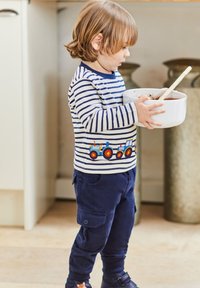 Dziecko w granatowych spodniach cargo i długim, w paski, kolorowym t-shircie z wzorem ciągnika, trzymające białą miskę z ciastem i drewnianą łyżką.