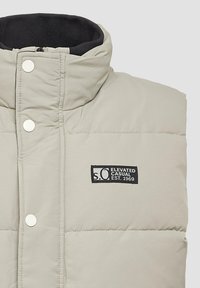 Jasno beżowa ocieplana kamizelka z wysokim kołnierzem, zatrzaskami i czarną naszywką z logo "Elevated Casual Est. 1969" na piersi.
