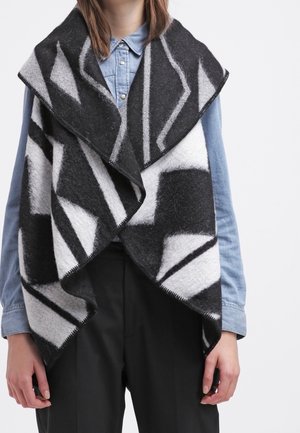 Woman wearing a black and white geometric patterned open-front vest over a blue button-up shirt with black trousers.