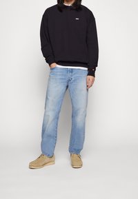 Svart sweatshirt med rund hals och subtil logotyp, ljusblå slitna jeans och tan mockaskor. Enkel, avslappnad outfit.