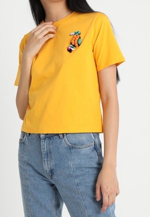 Kobieta w żółtej koszulce z kolorowym broszką w kształcie gąsienicy na piersi, w połączeniu z niebieskimi dżinsami.