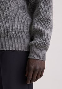 Maglione grigio lavorato a maglia con texture a coste. Presenta maniche lunghe e una vestibilità aderente ai polsini, abbinato a pantaloni scuri.
