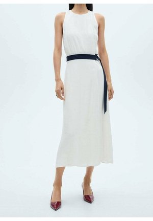 Vrouw draagt een witte mouwloze midijurk met een zwarte riem en rode puntige hakken, staand tegen een witte achtergrond.
