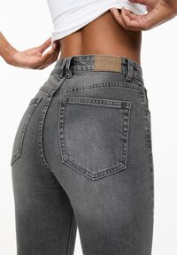 Jean en denim gris à coupe ajustée, avec une finition texturée, deux poches arrière et une étiquette en cuir beige sur la ceinture.