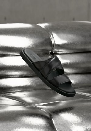 Zwarte slidesandaal met twee brede leren banden die boven een glanzende metalen gestoffeerde bank zweven.