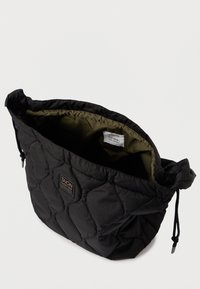 Μαύρη τσάντα ώμου με καπιτονέ σχέδιο και κλείσιμο με κορδόνι, εσωτερική κάλυψη σε ελιά, και ορθογώνια ετικέτα μάρκας στο εξωτερικό.