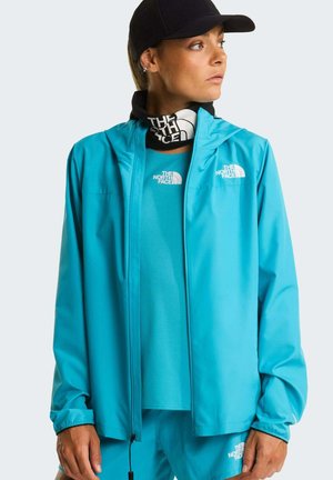 Vrouw draagt een felblauwe North Face-jas, bijpassend shirt en broek, een zwarte halswarmersjaal en een zwarte pet, kijkt opzij.