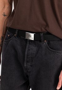 Cinturón negro trenzado con una hebilla de metal plateado que lleva un logo. Llevado sobre un jeans de mezclilla oscuros, mostrando una textura suave y un estilo casual.