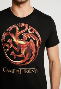 Schwarzes Baumwoll-T-Shirt mit einem lebhaften roten und goldenen Design eines dreiköpfigen Drachen und dem Text "Game of Thrones" am unteren Rand.