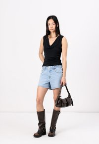 Top negro con cuello alto, pantalones cortos de mezclilla azul claro, botas marrones hasta la rodilla con hebillas y un pequeño bolso marrón. Fondo neutro.