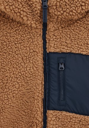 Tissu polaire brun et moelleux avec une surface texturée, panneau en nylon noir contrastant et détails en zip. Présente une fermeture à glissière sur toute la longueur.