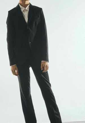 Traje negro a medida con una chaqueta ajustada y pantalones rectos, llevado sobre una camisa blanca con cuello, que cuenta con un solo botón y bolsillos.