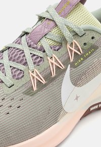 Buty do biegania w terenie Nike w jasnej zieleni i lawendowym odcieniu, z siateczkową cholewką, różowymi sznurowadłami, zaokrąglonym noskiem i teksturowaną podeszwą z białym logo Swoosh.