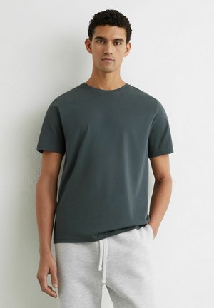 Młody mężczyzna z krótkimi kręconymi włosami, ubrany w ciemnoszary T-shirt z krótkim rękawem i jasnoszare spodnie dresowe, stojący na tle białej ściany.