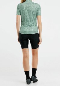 Maillot de cyclisme vert menthe à manches courtes avec des pois noirs, associé à un cuissard de cyclisme rembourré noir et des chaussures foncées.
