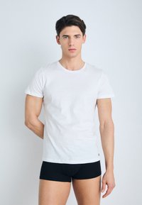 Camiseta de algodón blanca de manga corta, cuello redondo, textura suave, combinada con pantalones cortos ajustados negros, modelo de pie contra un fondo claro.