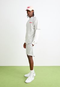 Chemise grise à manches courtes avec un logo rouge, shorts gris, baskets blanches et une casquette rose clair. En fond, un sol vert uni.