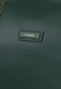 Donkergroene tas met een gladde, matte textuur. Beschikt over een rechthoekig label met "HEXAGONA" in het wit gedrukt op een zwarte achtergrond.