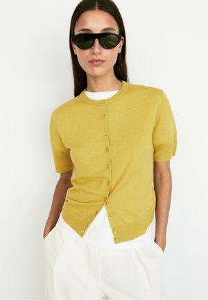 Femme portant des lunettes de soleil noires, un cardigan à manches courtes jaune moutarde boutonné sur une chemise blanche, et un pantalon blanc avec les mains dans les poches.