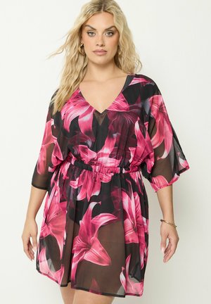 Femme blonde portant une robe noire transparente avec de grandes impressions florales roses et des manches trois-quarts, debout devant un fond uni.