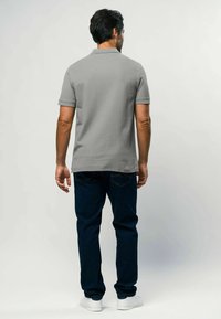 Vīrietis stāv ar muguru pret mums, tērpies pelēkā īsroku polo krekliņā, tumši zilās džinsās un baltos sporta apavos uz vienkārša fona.