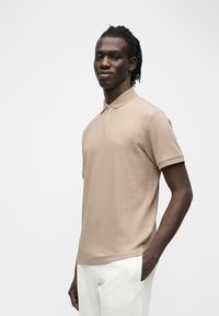 Homme aux dreadlocks courtes portant un polo beige côtelé à manches courtes et un pantalon blanc, une main dans la poche, sur un fond clair uni.