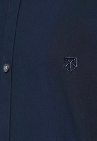 Camicia blu navy con una texture liscia, caratterizzata da un piccolo logo ricamato a forma di scudo sul lato sinistro del petto. Dettagli dei bottoni visibili.