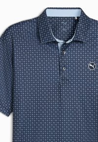 Sininen polo-paita, jossa on kuvio pienistä ympyröistä, vaaleansininen kauluksen ja logo vasemmassa rinnassa.