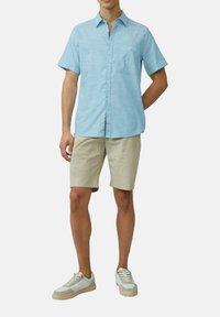 Camicia azzurra a maniche corte con bottoni, caratterizzata da un motivo tessurizzato e una tasca sul petto, abbinata a pantaloni corti beige e sneaker bianche.