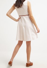 Vestido beige con lunares, con un corsé ajustado y una falda acampanada. Presenta un cinturón marrón ceñido en la cintura y una textura de tela suave.