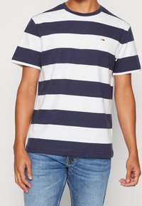 Camiseta de manga corta a rayas en azul marino y blanco, con cuello redondo, fabricada en algodón. Presenta un pequeño logo en el pecho y detalles de dobladillo cosidos.