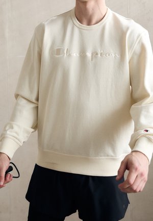 Person iført en cremefarvet Champion sweatshirt med logo på bryst og ærme, og sorte shorts, holder sorte briller i venstre hånd.