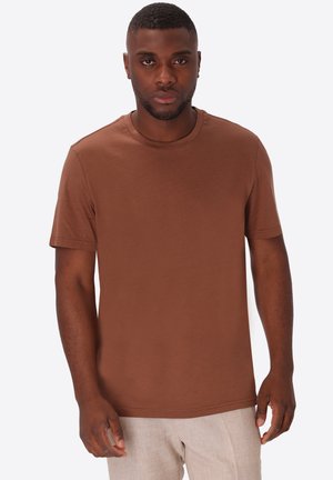 Uomo con capelli corti e barba, che indossa una maglietta marrone tinta unita a maniche corte e pantaloni beige chiaro, in piedi davanti a uno sfondo bianco.