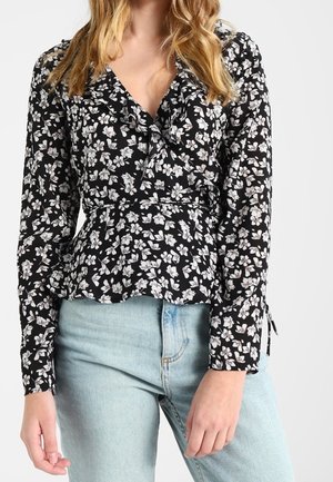 Kobieta ubrana w czarną bluzkę z długim rękawem w biały kwiatowy wzór oraz jasnoniebieskie, wysokie dżinsy na jednolitym tle.