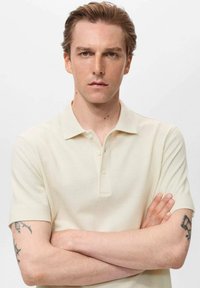 Tricou polo de culoare bej deschis, dintr-un material texturat, având guler și un sistem de închidere cu trei nasturi, mâneci scurte și o croială lejeră.