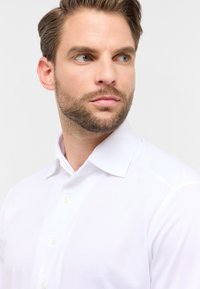 Witte overhemd met een gladde textuur, voorzien van een voorflap en vier knopen. Het model heeft een korte baard en gestyled haar.