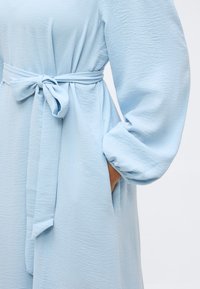 Φόρεμα από ανοιχτό μπλε υφασμάτινο με υφή, με μακριά μπαλόν μανίκια και ζώνη στη μέση που δένει, χέρι τοποθετημένο μέσα σε πλαϊνή τσέπη.