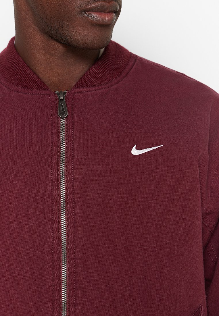 Uomo che indossa una giacca a zip color mogano con il logo Nike bianco sul petto, inquadrato dal collo alla parte centrale del torso.