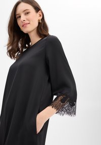 Zwarte satijnen jurk met driekwartmouwen afgewerkt met fijn zwart kanten randje, rechte snit en zijzakken. Overal een gladde textuur.