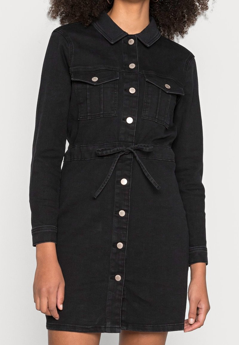 Femme portant une robe chemise en jean noire avec des poches avant et une ceinture à la taille, montrée des épaules jusqu'à mi-cuisse.