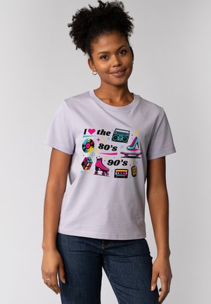 Jonge vrouw die glimlacht, gekleed in een lichtpaars t-shirt met kleurrijke iconen en teksten uit de jaren 80 en 90, staand voor een effen grijze achtergrond.