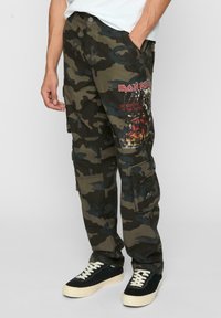 Kamo nohavice v tmavozelenej a čiernej farbe, ozdobené grafickým potlačou Iron Maiden, viacerými vreckami a voľným strihom.